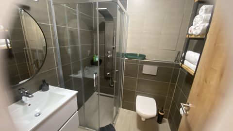 Celá koupelna: prosklený sprchový kout, umyvadlo se skříňkou, kulaté zrcadlo, závěsné WC a police s ručníky